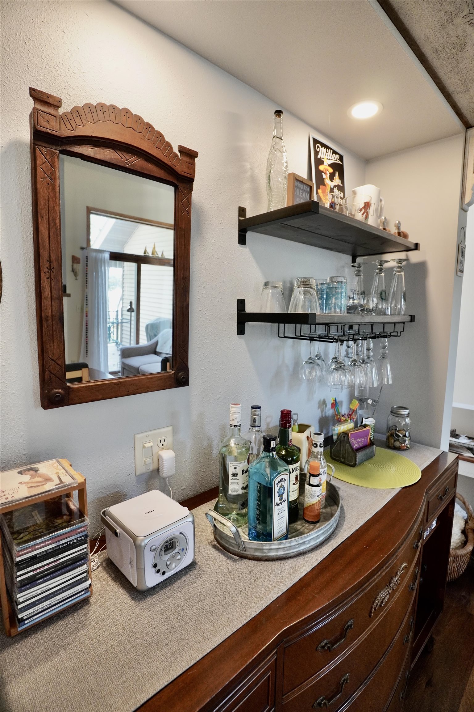 Furnished home bar with liquor bottles, hanging glassware, black shelves, and an ornate wooden mirror.