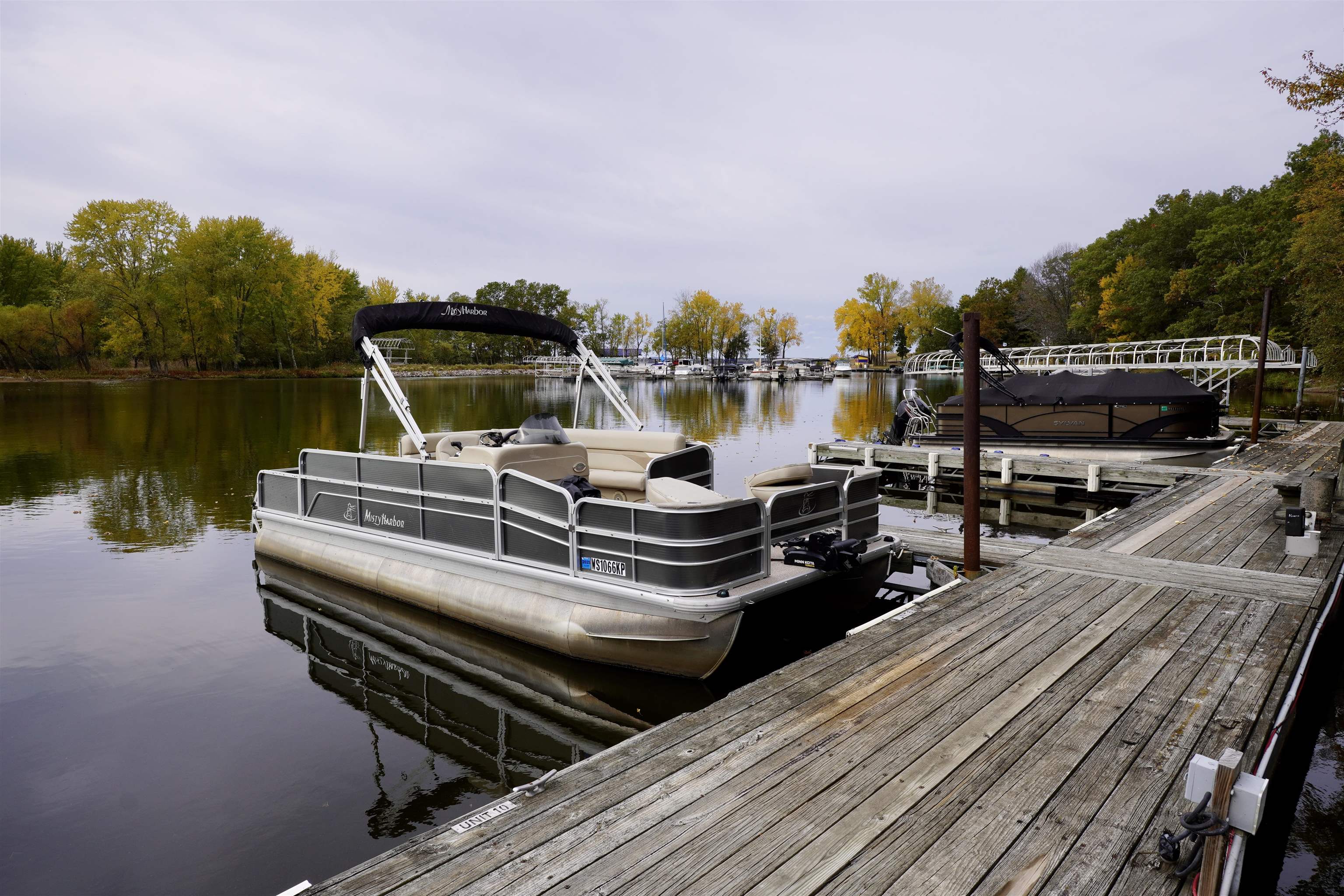 Pontoon boat moored beside a wooden marina dock, with autumn trees and boats across the water.