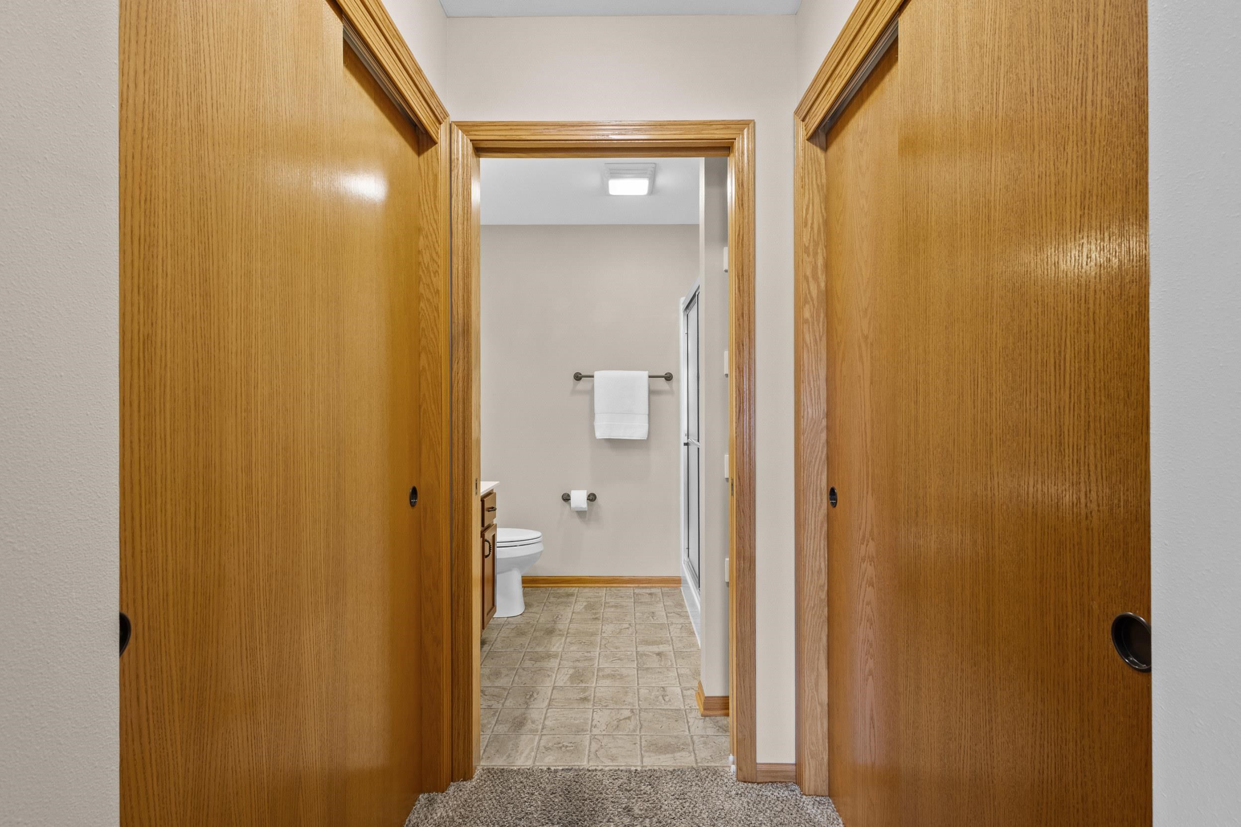 Bathroom viewed through open wooden doors, with beige tile flooring and a glass shower.