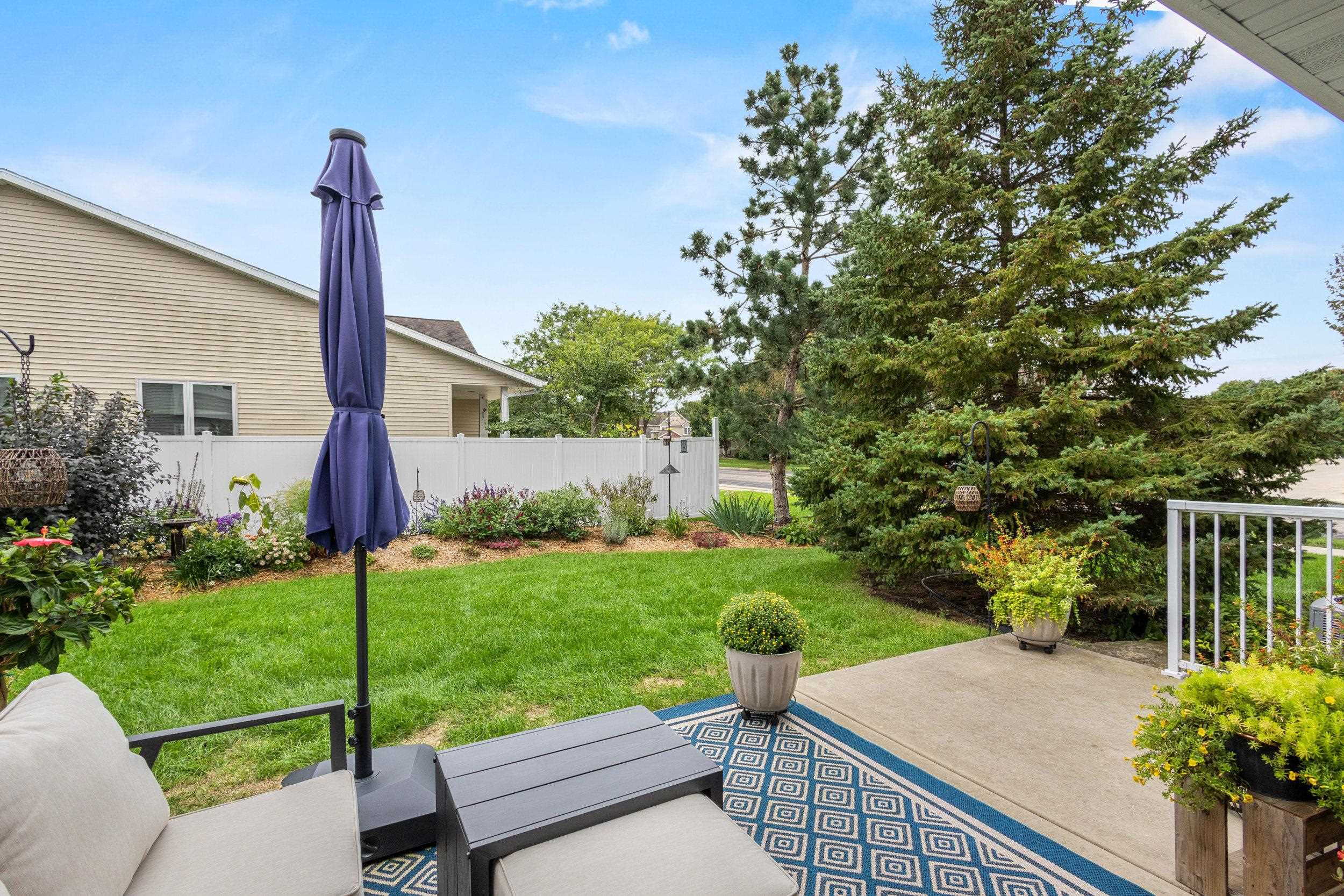 Backyard patio with cushioned seating and a purple umbrella overlooking a landscaped lawn and evergreen trees.