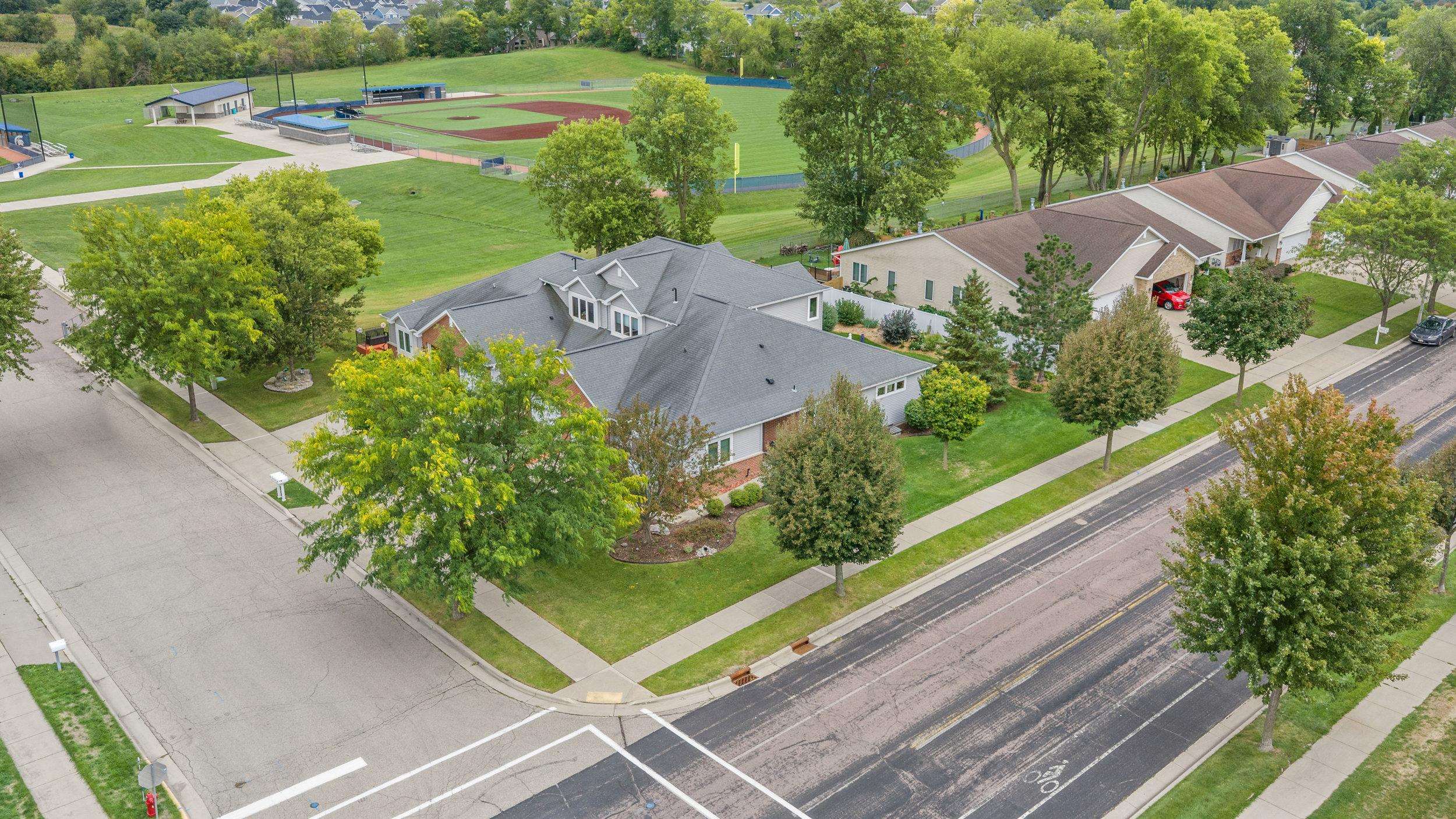 Gray-roofed home at a street corner beside a tree-lined neighborhood and baseball field.