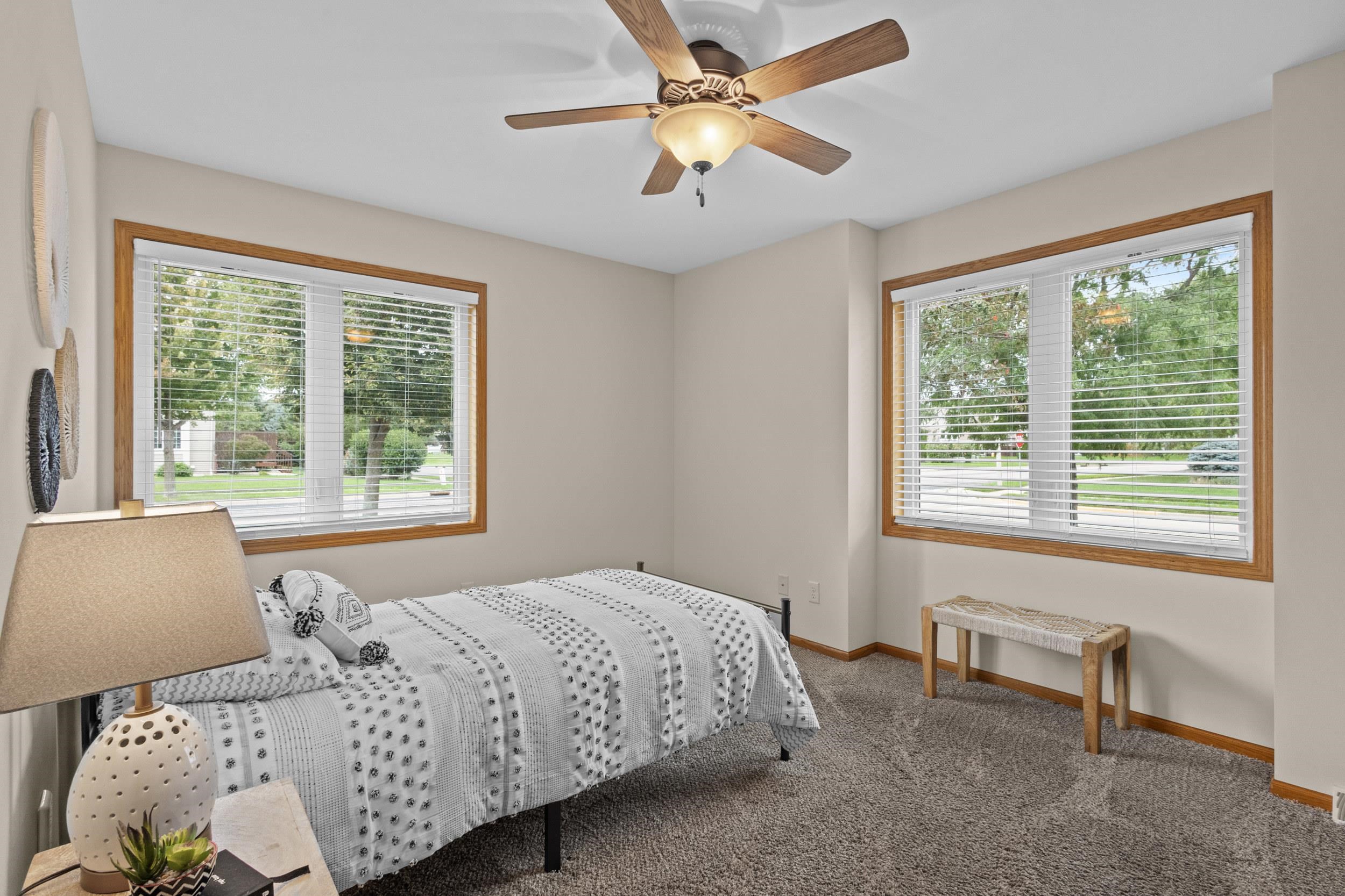 Bedroom with patterned white bedding, two wood-trimmed windows, gray carpet, and a ceiling fan.