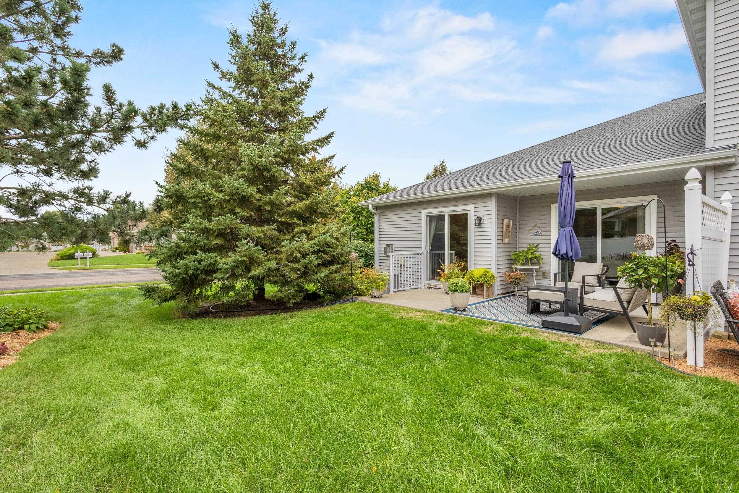 Home exterior with a furnished patio, purple umbrella, potted plants, and mature evergreens beside a broad lawn.