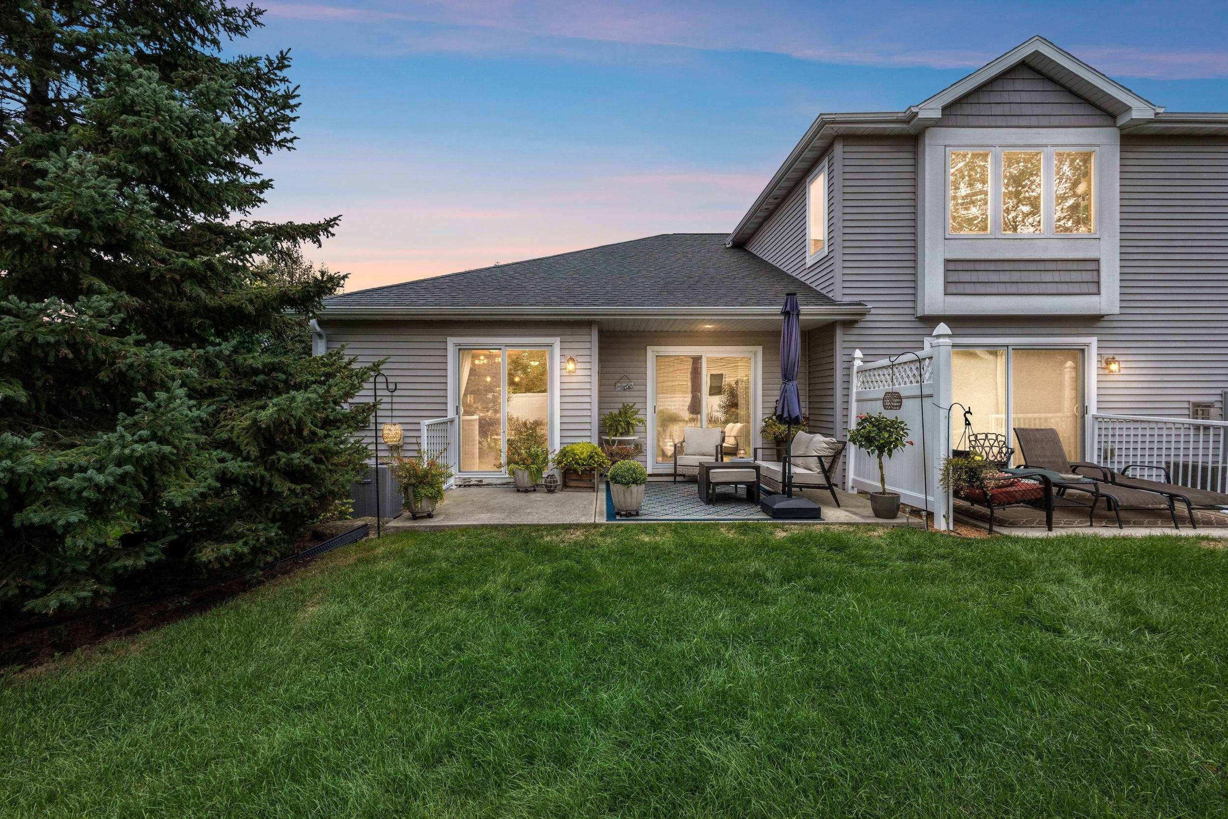 Rear of a gray two-story house with a furnished patio, privacy screen, and broad lawn at dusk.