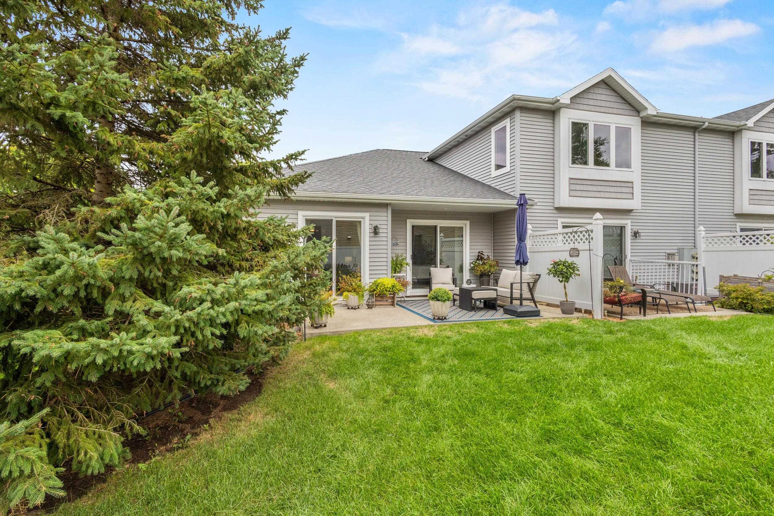 Gray two-story home with a furnished patio, white fencing, and a large evergreen beside the lawn.