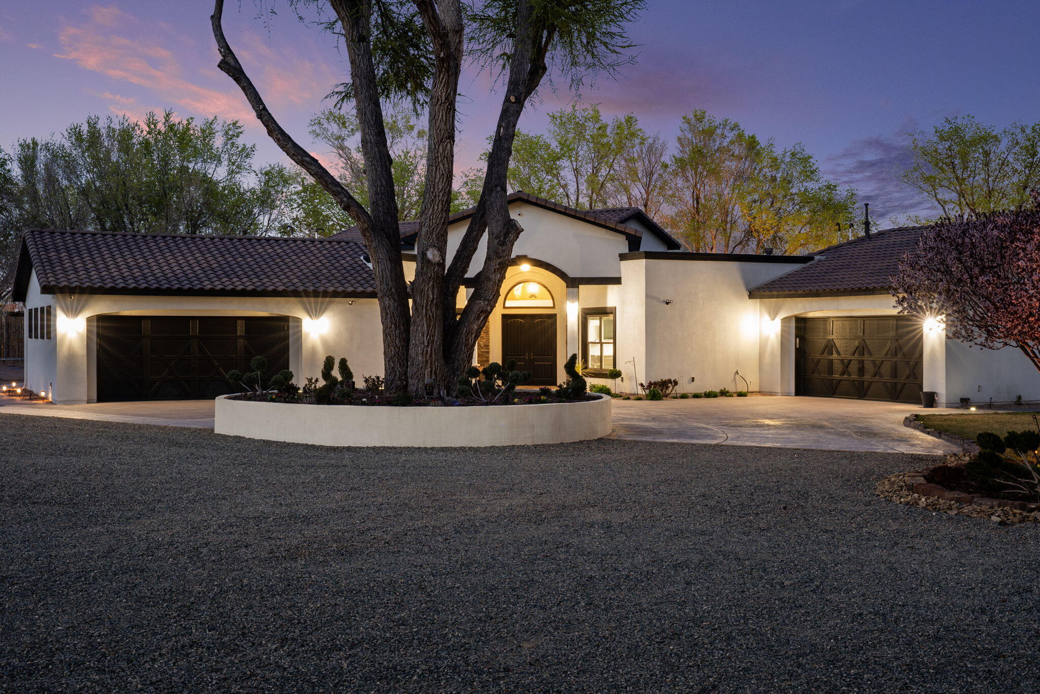 Stucco home at dusk with two garages and a large tree in a circular planter.