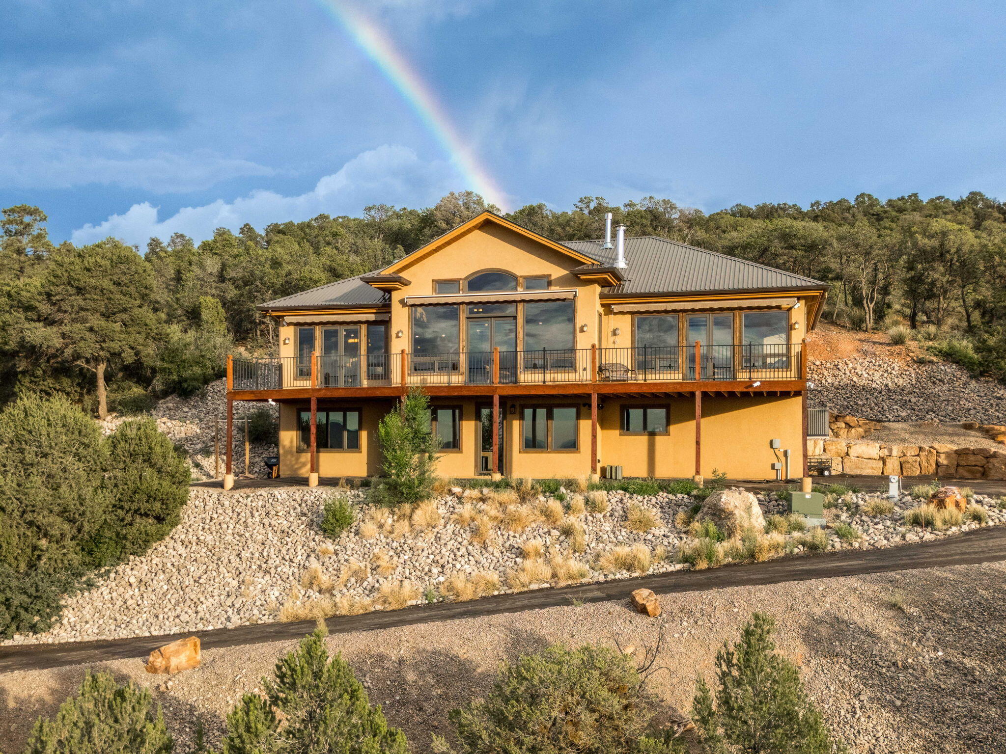 Ochre hillside home with a long deck beneath a rainbow.
