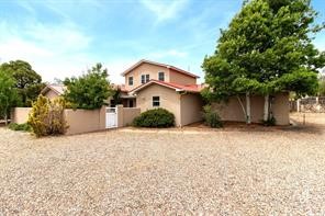Two-story tan home with a reddish roof, gravel yard, and mature trees.