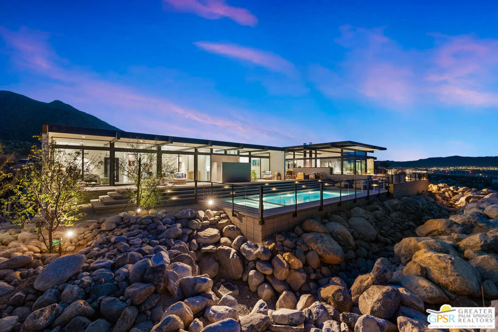 Modern glass home and illuminated pool among desert boulders, with mountains behind at dusk.