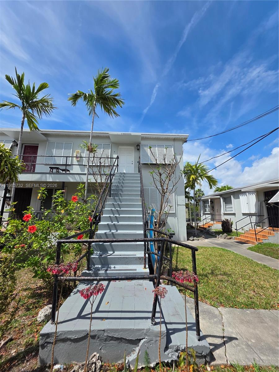 Light-gray two-story residence with an outdoor staircase, palm trees, and flowering plants.