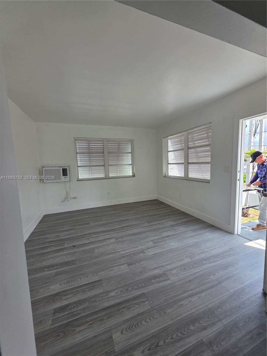 Empty room with gray plank flooring, two blind-covered windows, and a wall-mounted air conditioner.