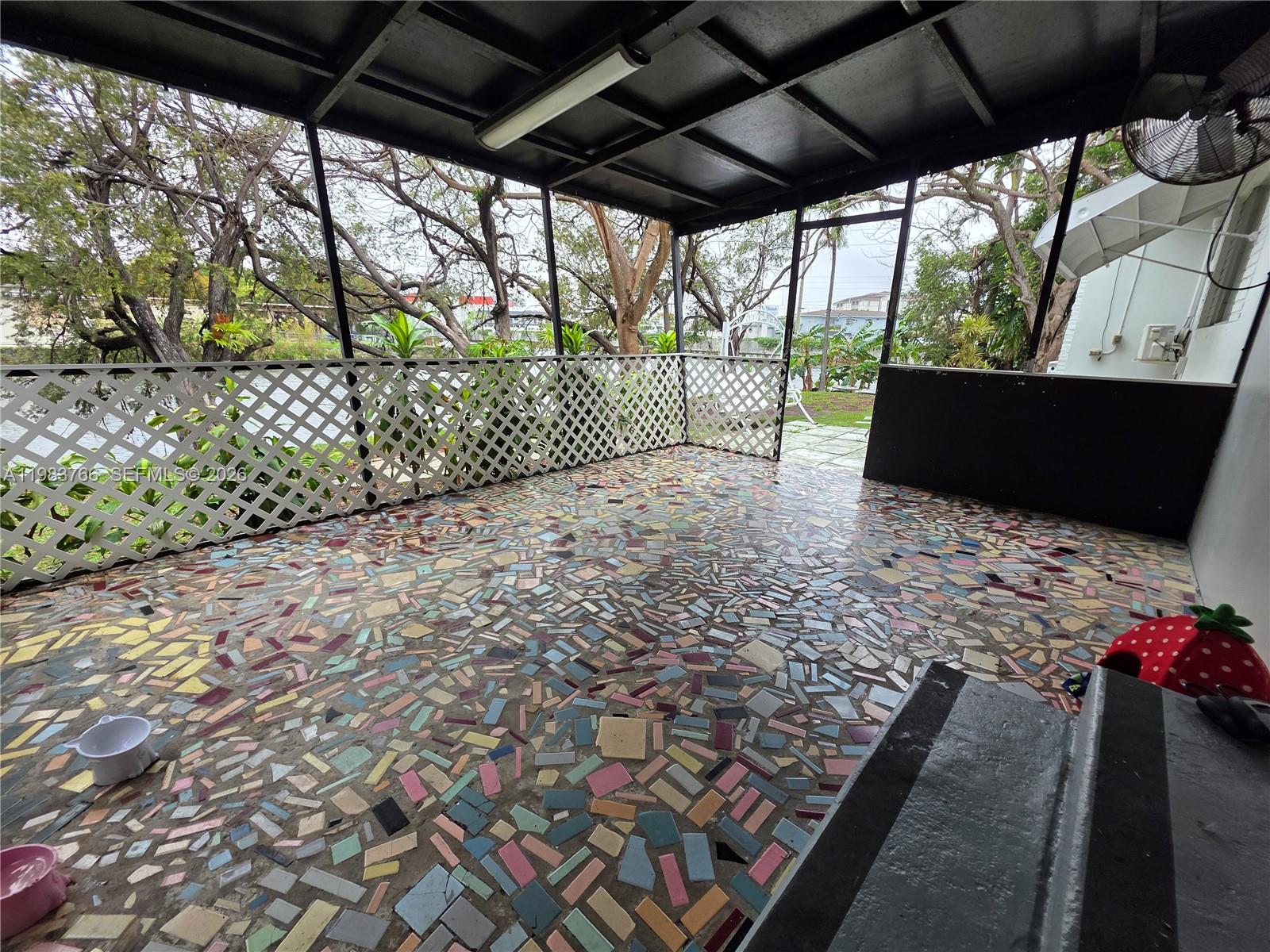 Covered patio with a colorful mosaic tile floor and white lattice railing overlooking a leafy garden.