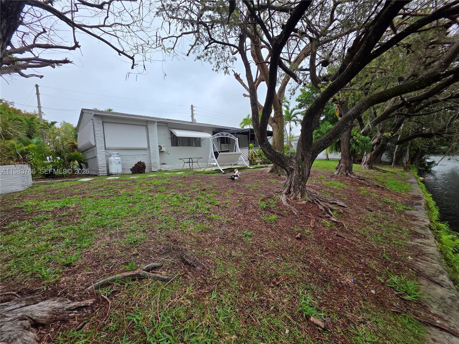 Single-story white house beneath mature trees, with an outdoor swing and canal beside the yard.