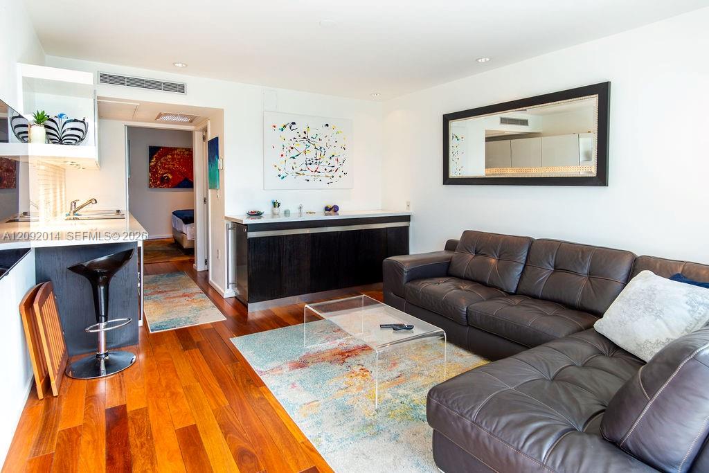 Living room with a dark leather sectional, colorful rug, and hardwood floors beside the kitchen.