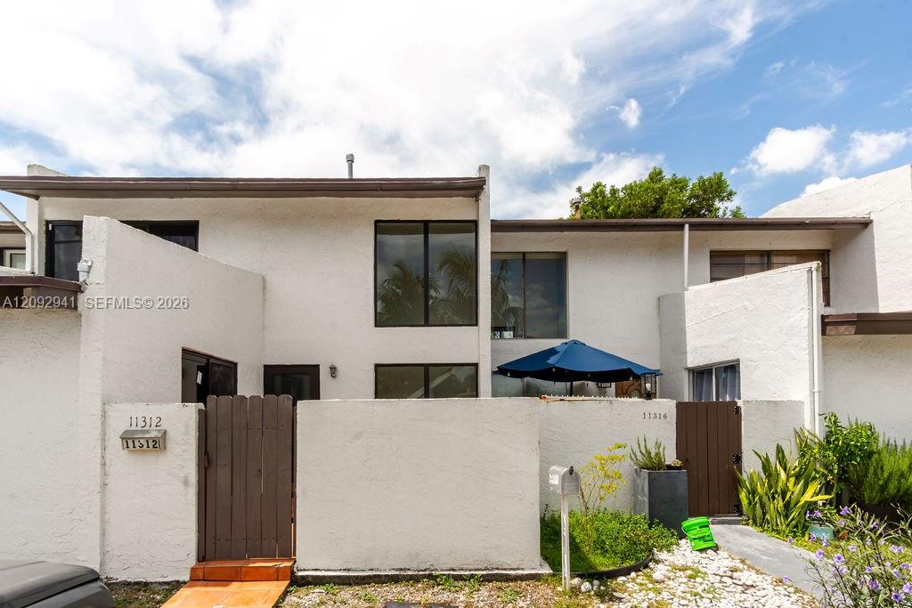 White two-story residence with a walled courtyard, brown gates, and a blue patio umbrella.