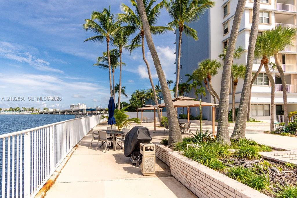 Waterfront condo patio with outdoor seating, a covered grill, and palm trees beside the water.