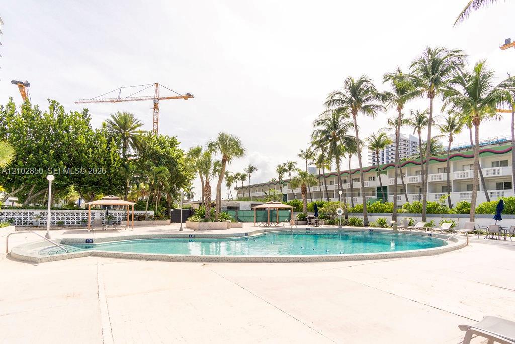 Outdoor swimming pool with palm trees, lounge chairs, and a residential building behind it.
