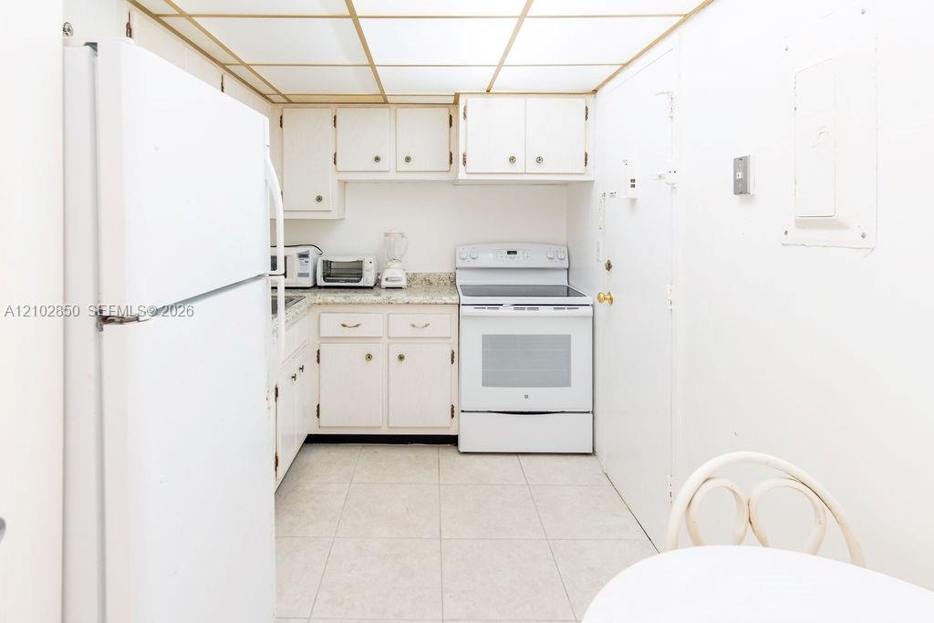 Narrow white kitchen with a large refrigerator, electric range, and tiled floor.