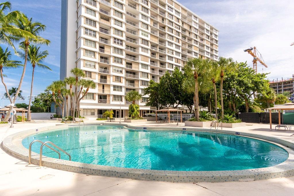 Condominium building behind a turquoise pool, framed by palm trees.