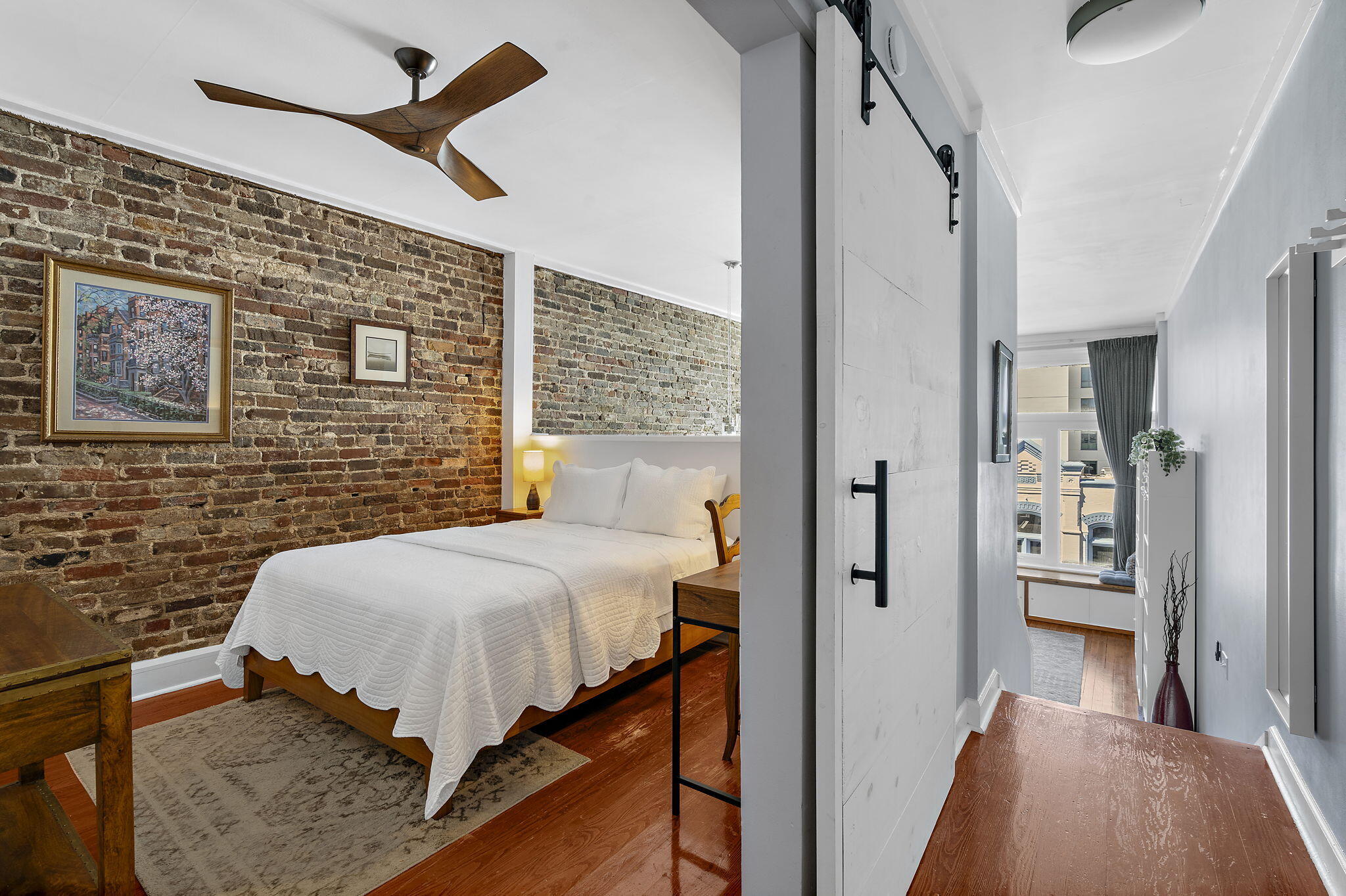 Furnished bedroom with an exposed brick wall, white bed, and view of nearby buildings through the window.