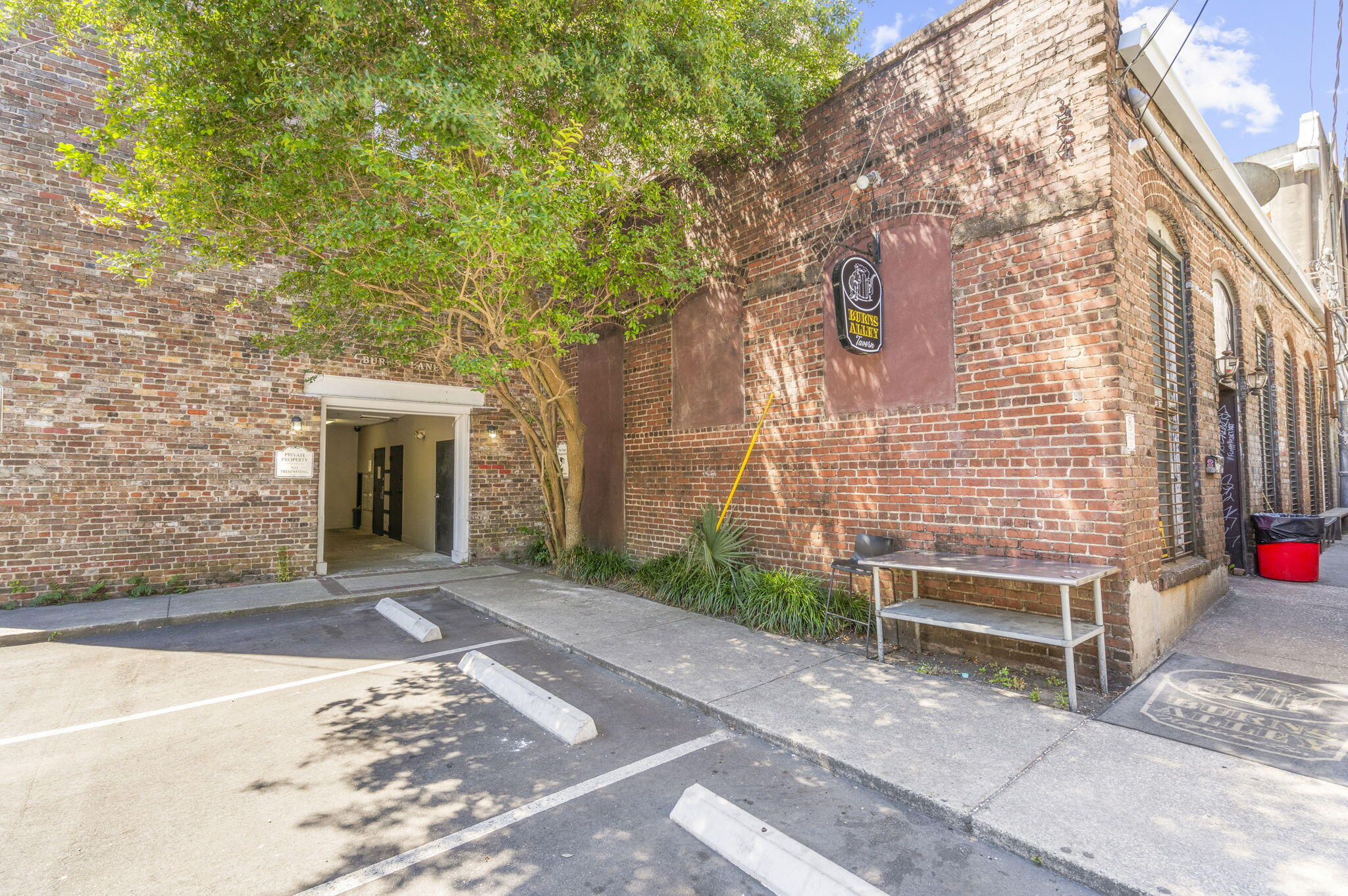 Brick building entrance beneath a leafy tree, beside a Burns Alley Tavern sign and small parking area.