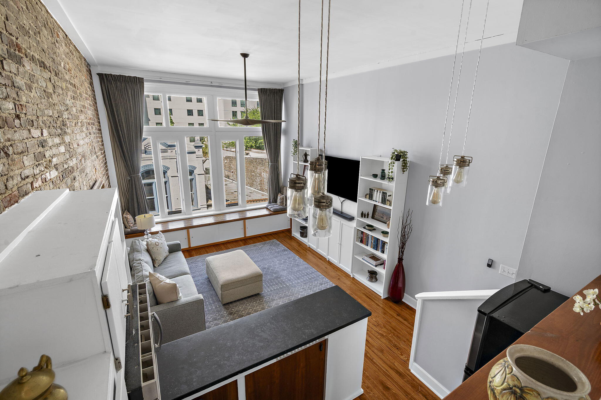 Open-plan living room with exposed brick, tall windows, and a built-in window seat.
