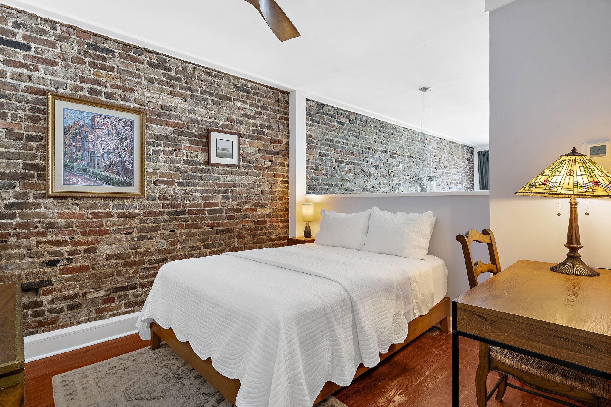 Bedroom with a white bed, exposed brick walls, and a wooden desk with stained-glass lamp.