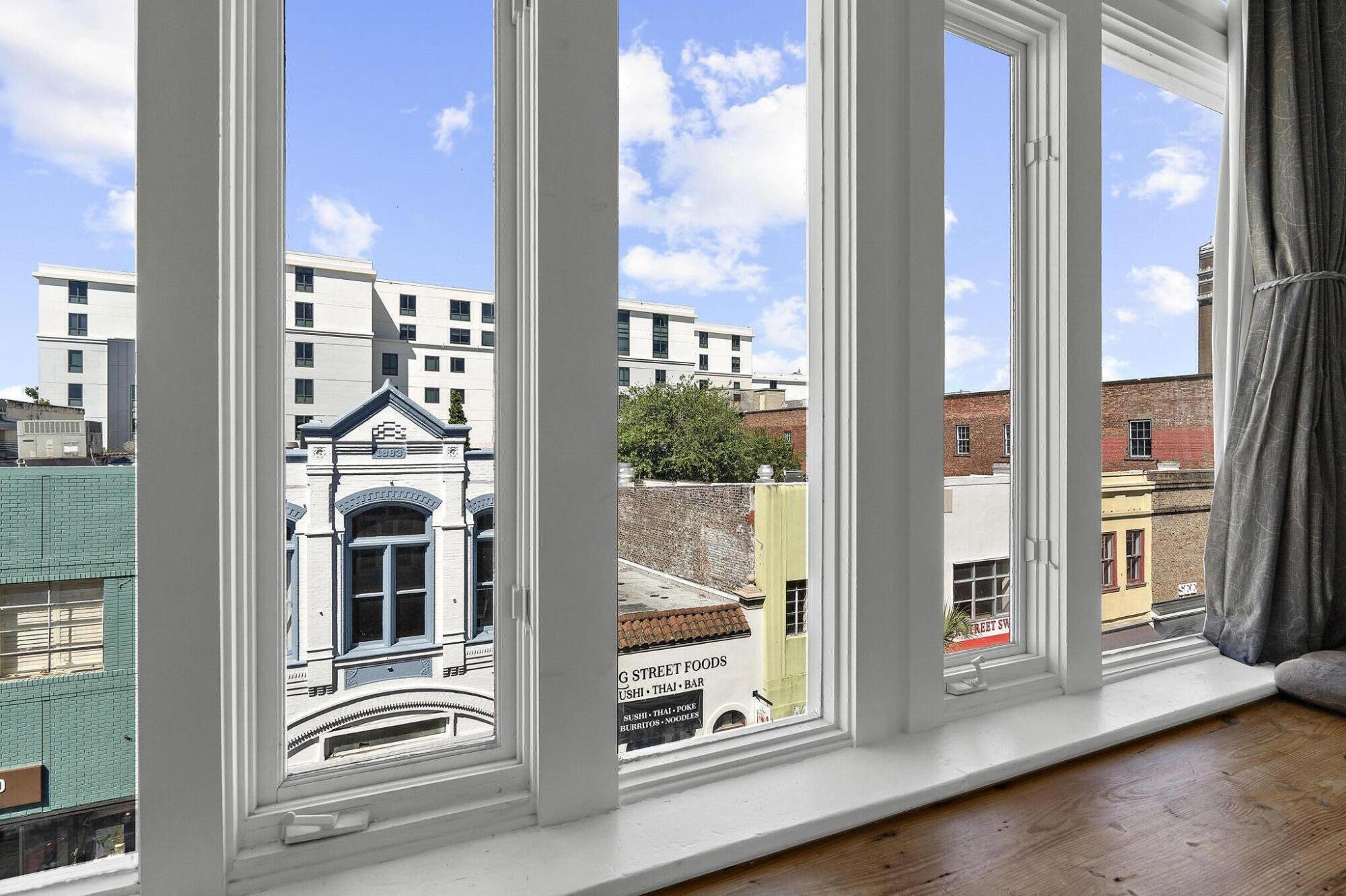 Charleston city street view through tall apartment windows, framed by white trim and a hardwood floor.