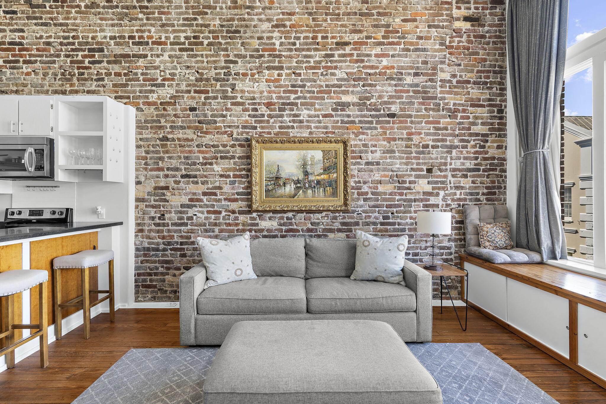 Living room with exposed brick wall, gray sofa, and a window seat overlooking nearby buildings.