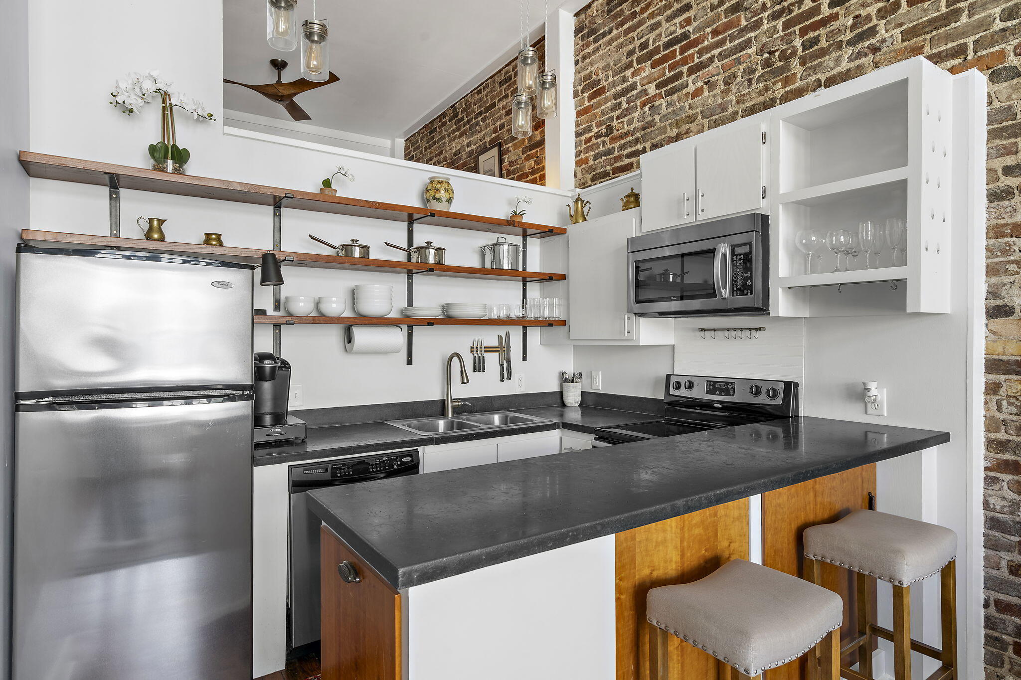 Kitchen with a large breakfast bar, stainless steel appliances, open shelves, and an exposed brick wall.