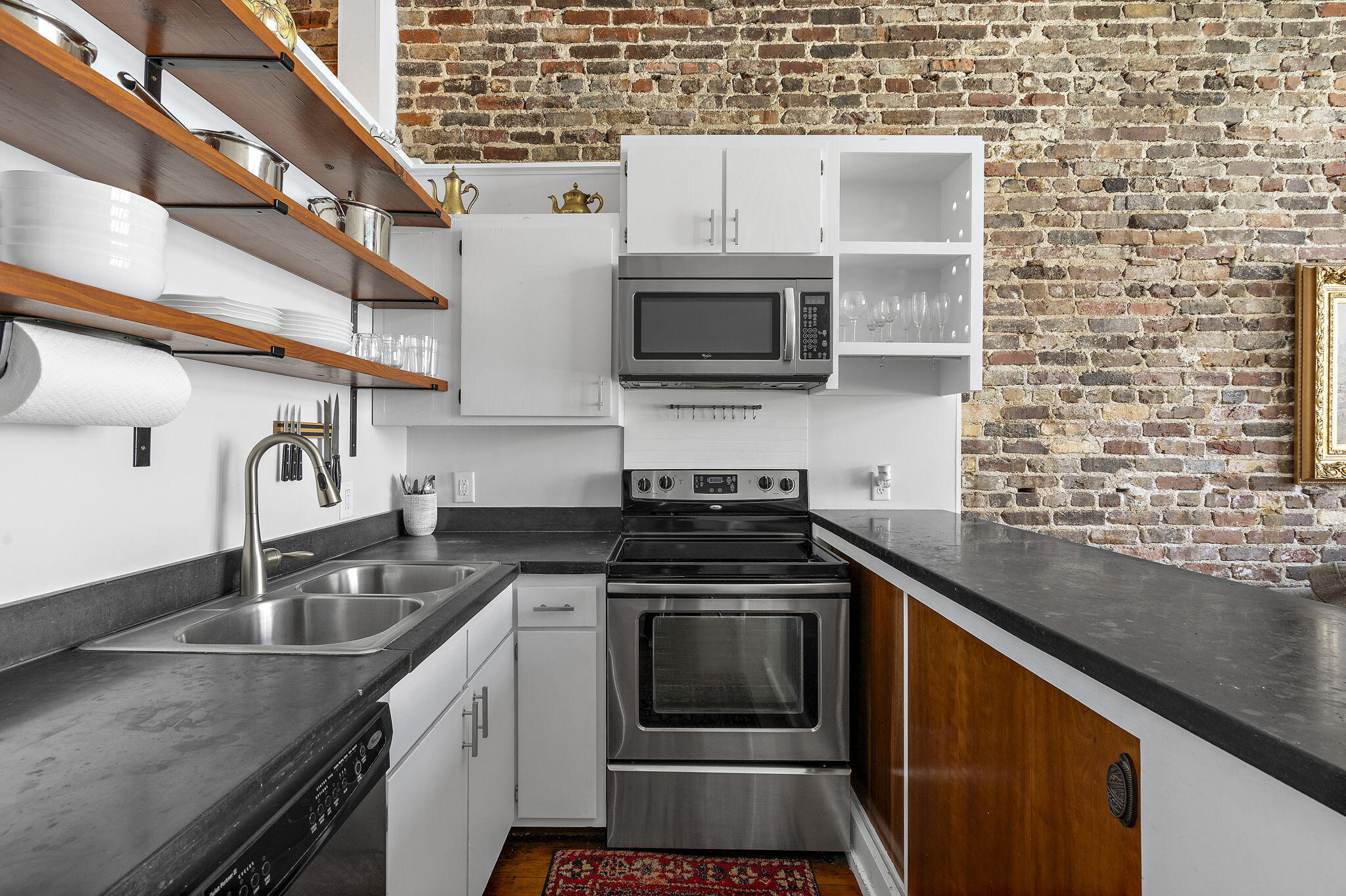 Compact kitchen with exposed brick, open wood shelves, dark counters, and stainless steel appliances.