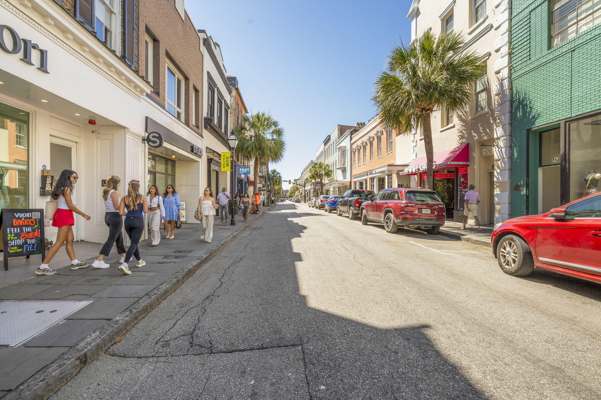 King Street storefronts and palm trees line a sunny Charleston street with pedestrians and parked cars.