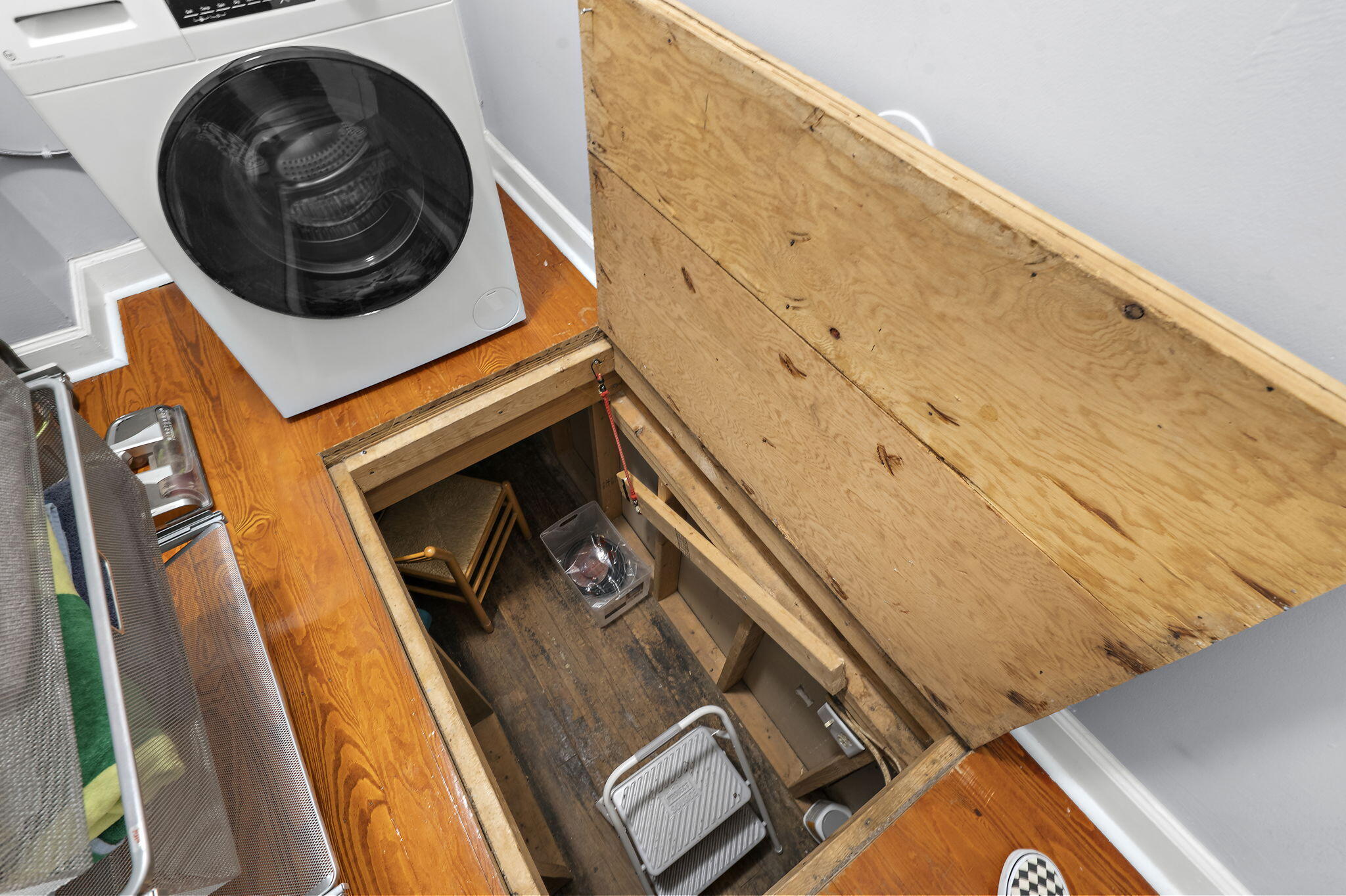 Laundry area with a front-loading washer beside an open floor hatch to a lower level.