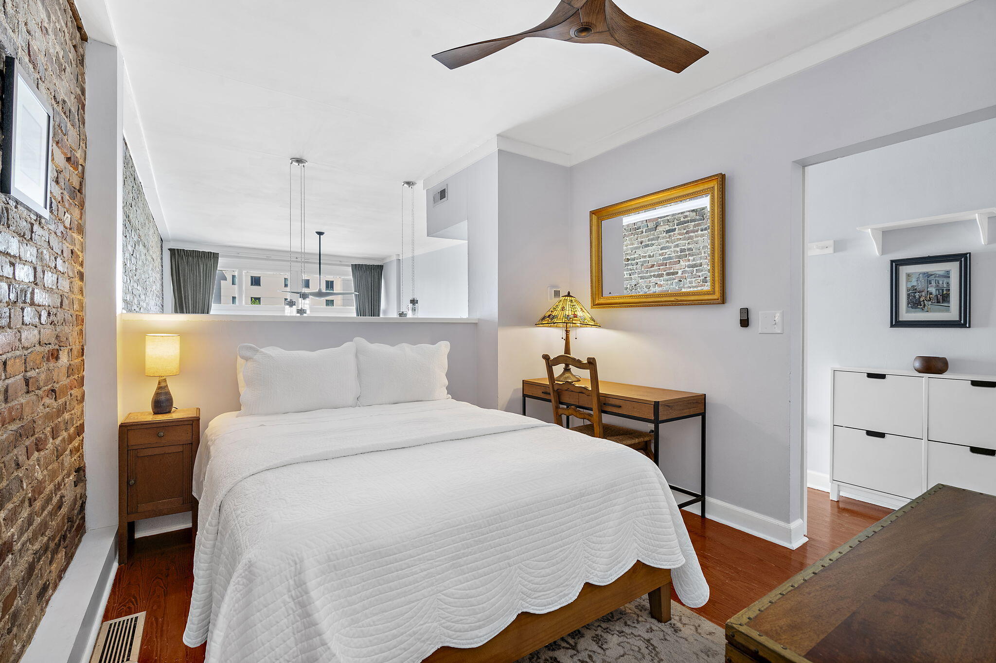 Bedroom with a white bed, exposed brick wall, hardwood floors, and a wooden desk.