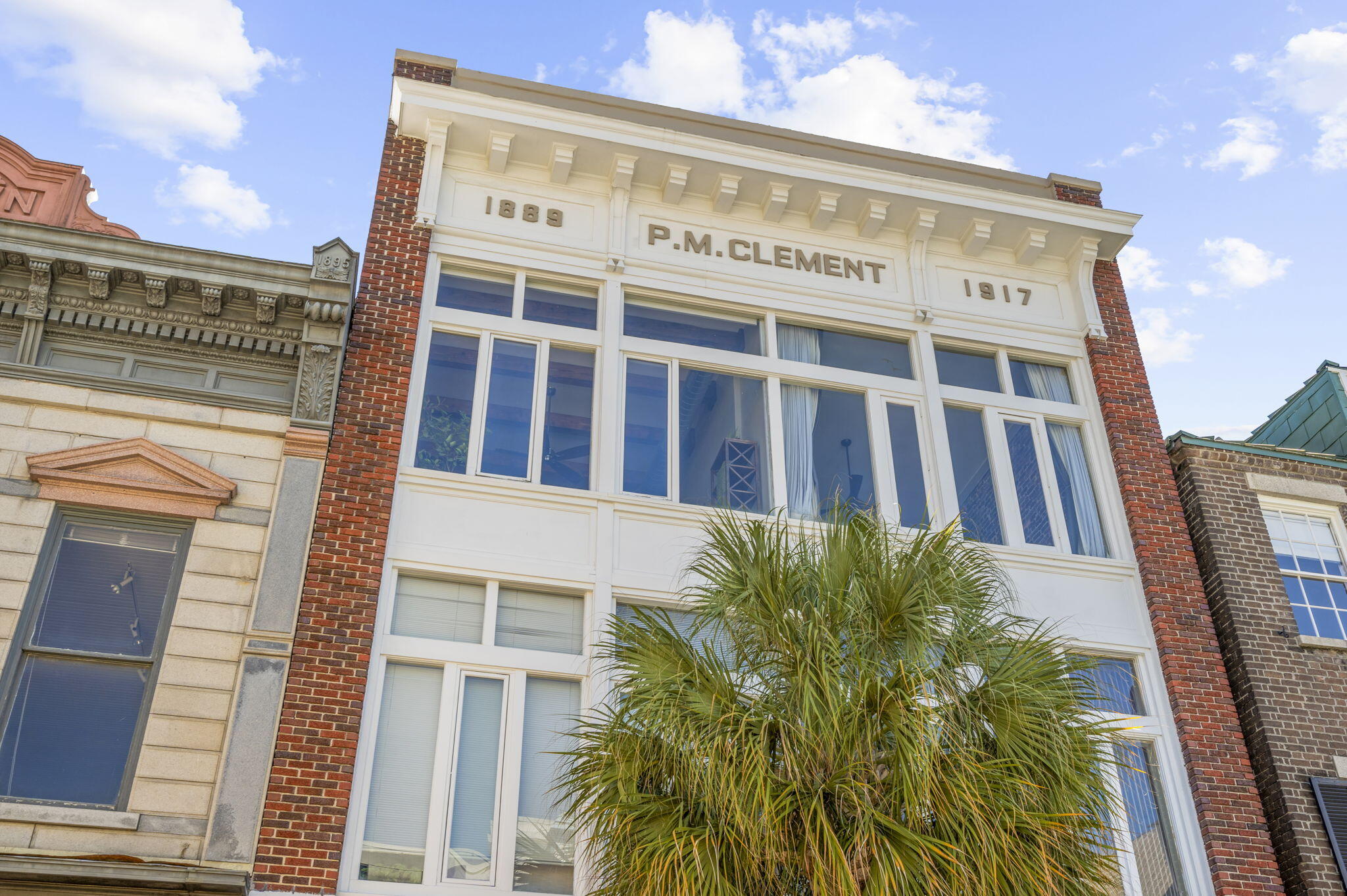 Historic brick building with white-trimmed windows, P.M. CLEMENT lettering, and a palm tree in front.