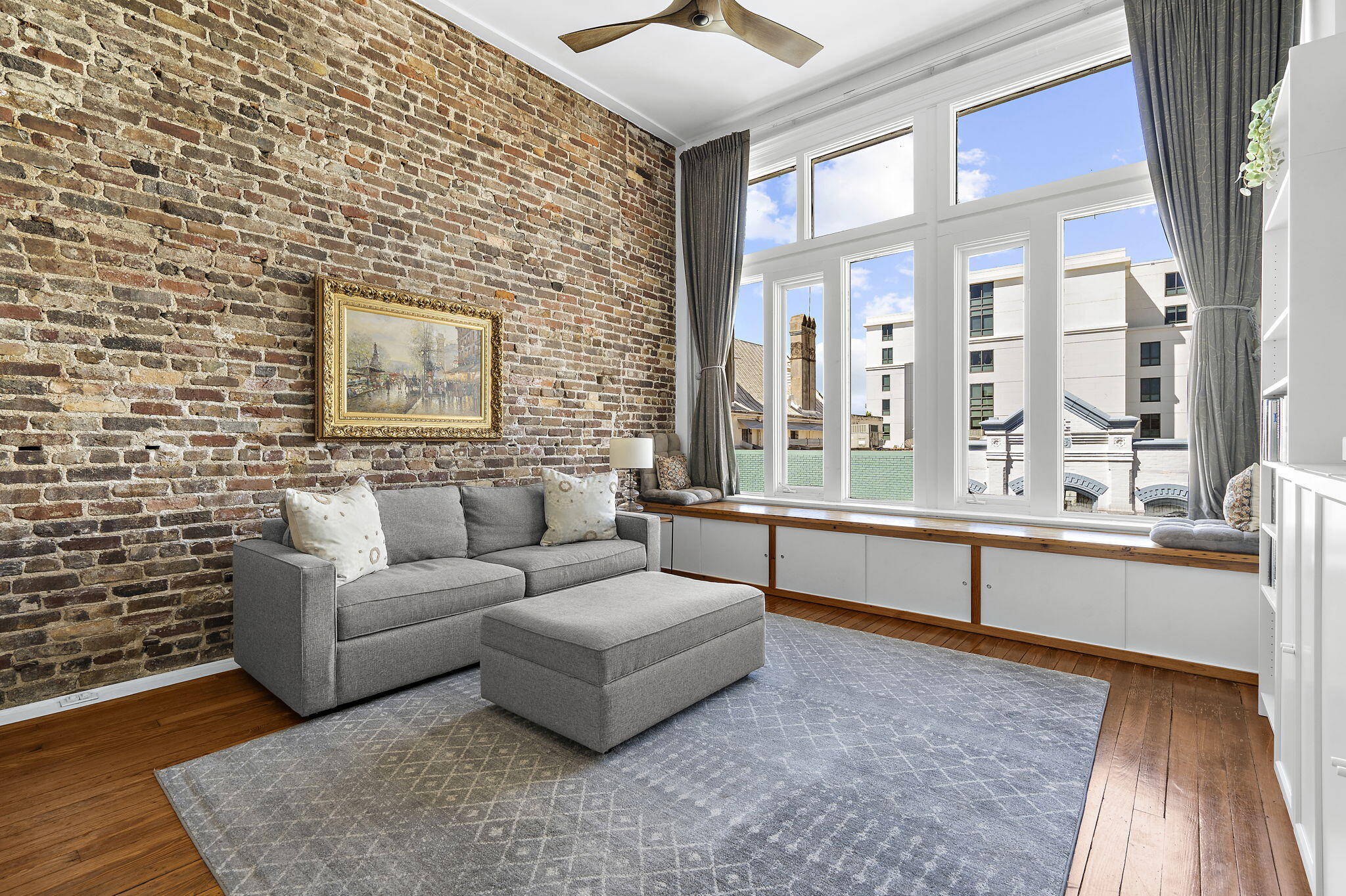 Living room with an exposed brick wall, gray sofa, hardwood floors, and tall windows.