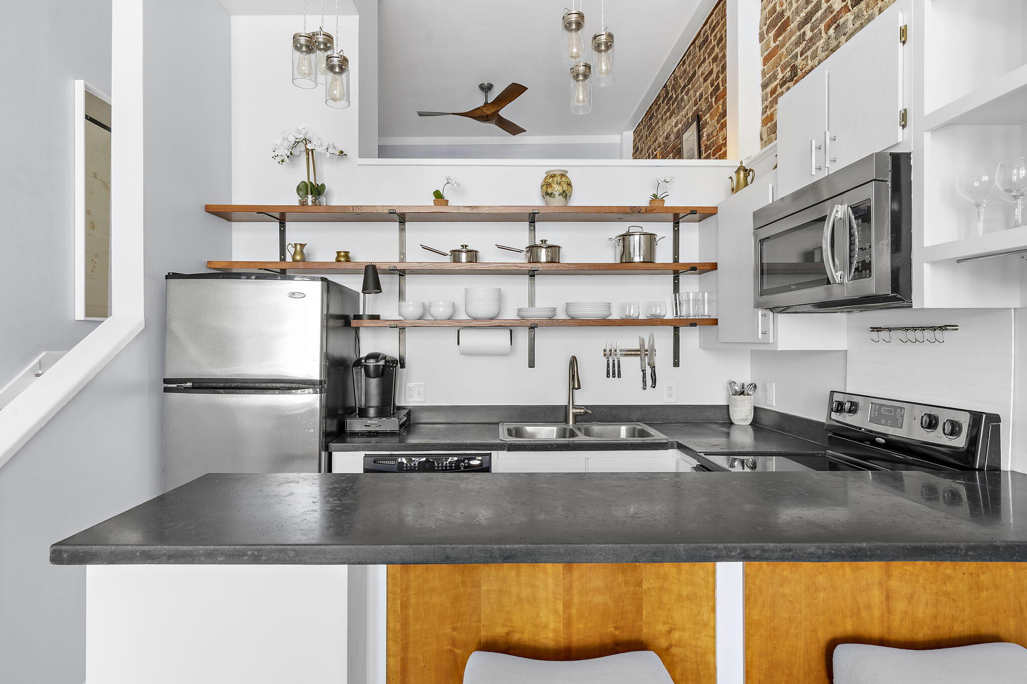 Modern kitchen with stainless steel appliances, open wooden shelving, and a broad dark-stone island.