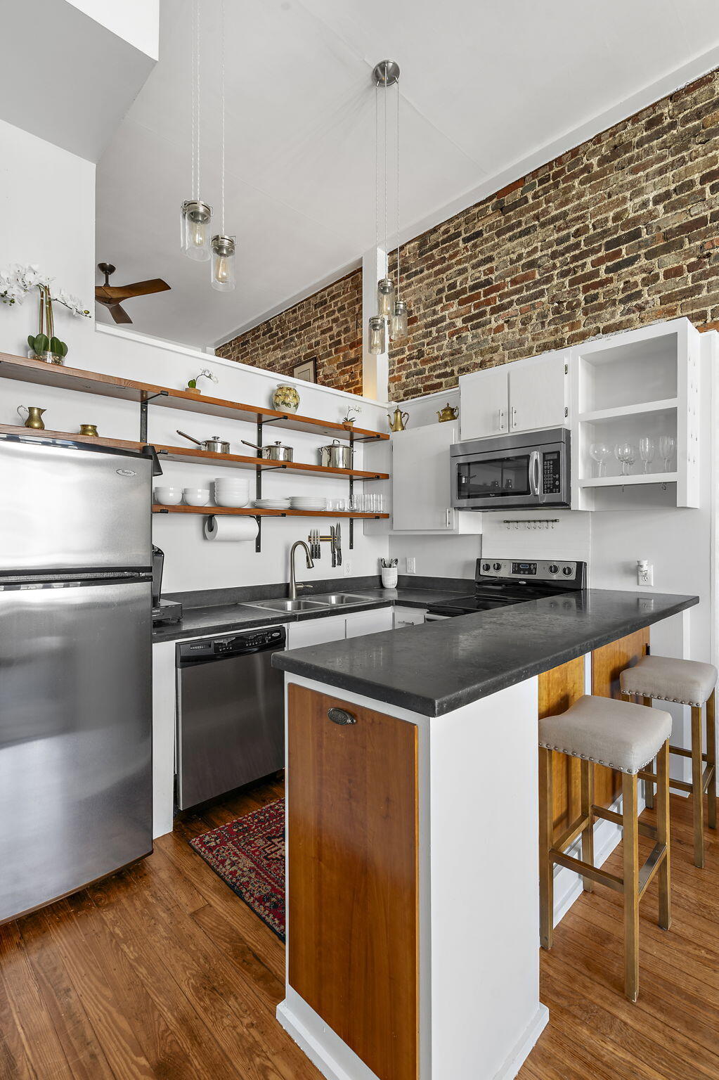 Loft-style kitchen with exposed brick, open wood shelving, stainless appliances, and a stone island with bar seating.