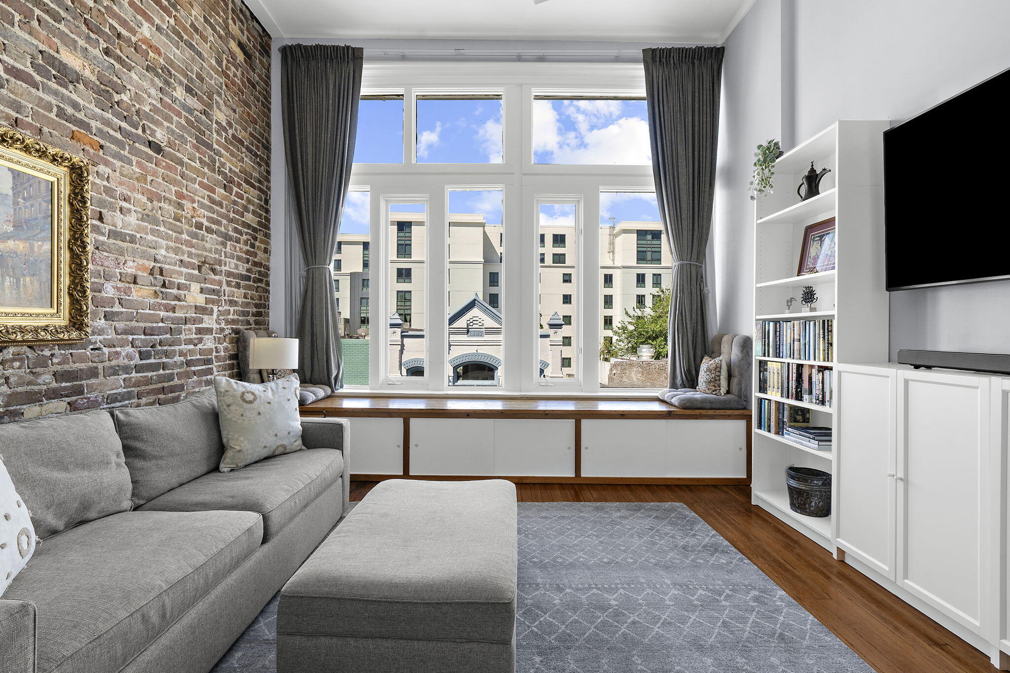 Living room with an exposed brick wall, gray sectional, and tall windows above a built-in window seat.
