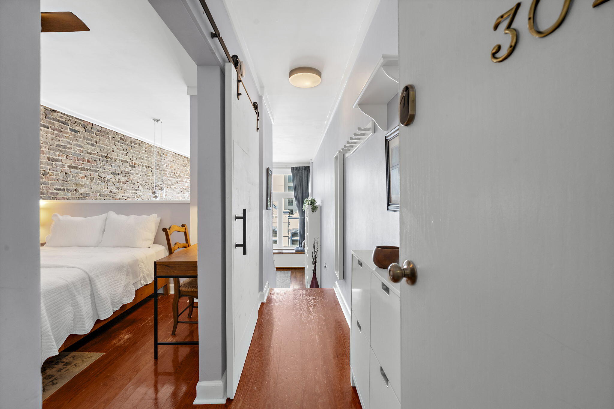 Furnished bedroom with an exposed brick wall and white bed beside a narrow hallway.