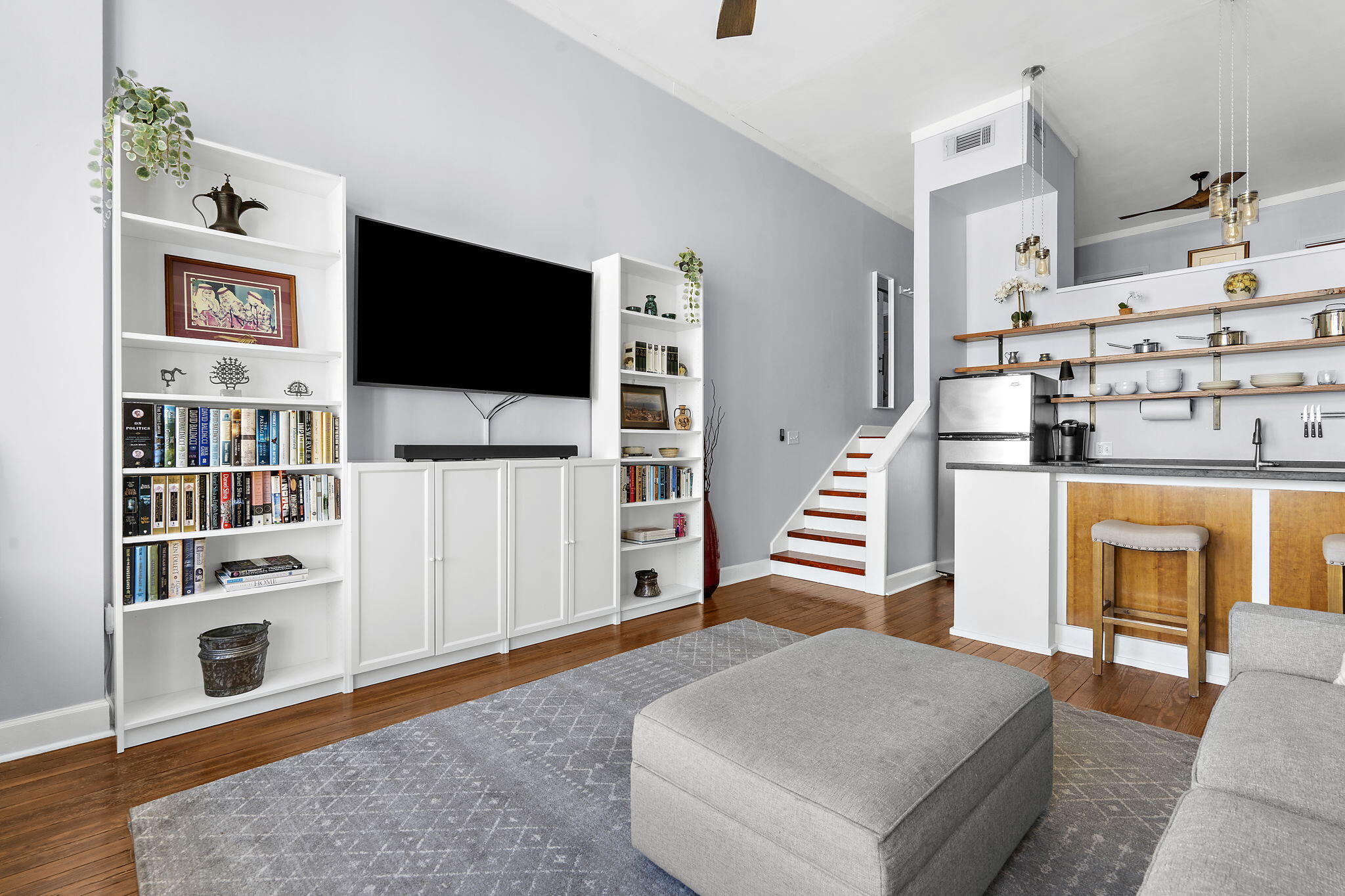 Open-plan living room and kitchen with built-in bookcases, gray seating, hardwood floors, and stairs.