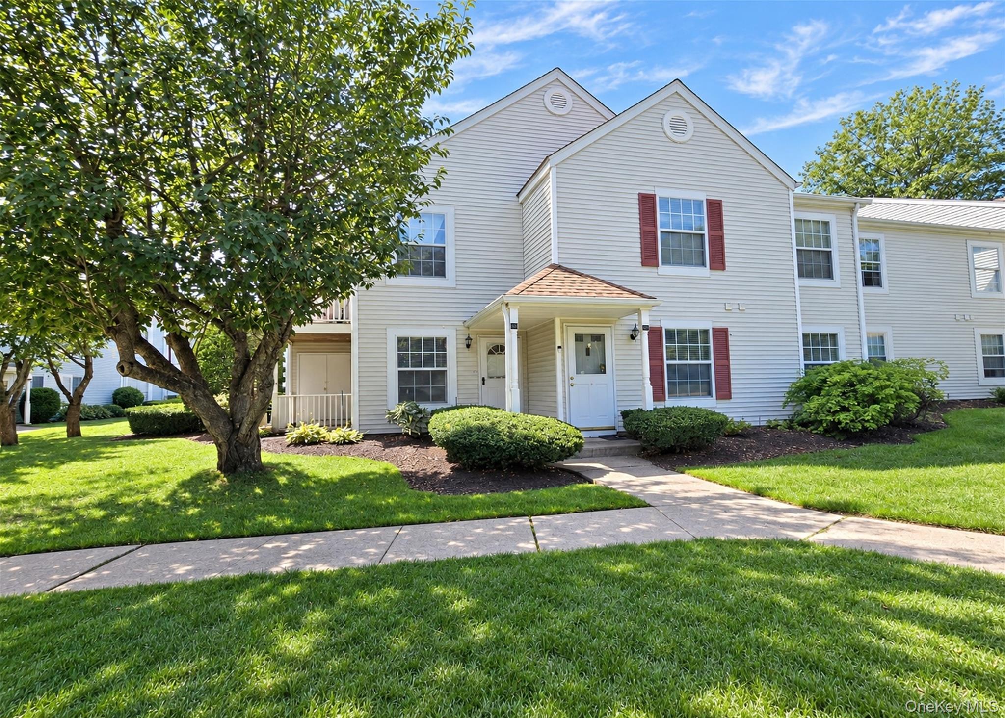 White two-story attached home with red shutters, shaded by mature trees beside a green lawn.