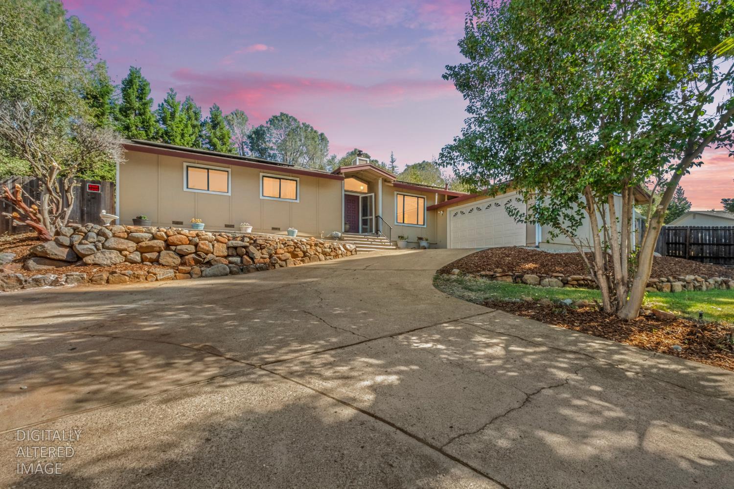 Single-story beige home with an attached garage, broad driveway, stone retaining wall, and pink dusk sky.
