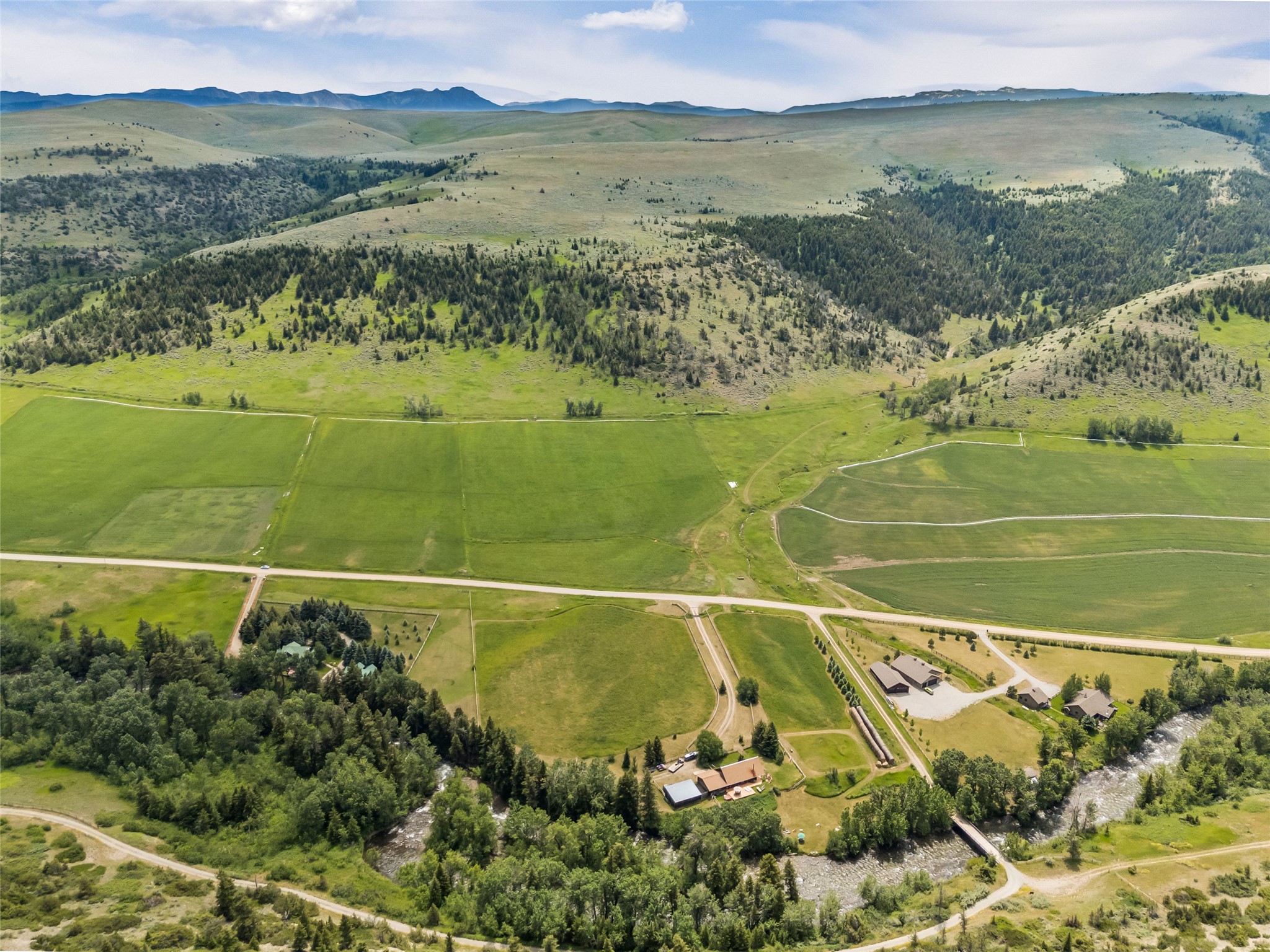 Aerial view of green fields, homes, and a tree-lined stream below rolling mountain foothills.