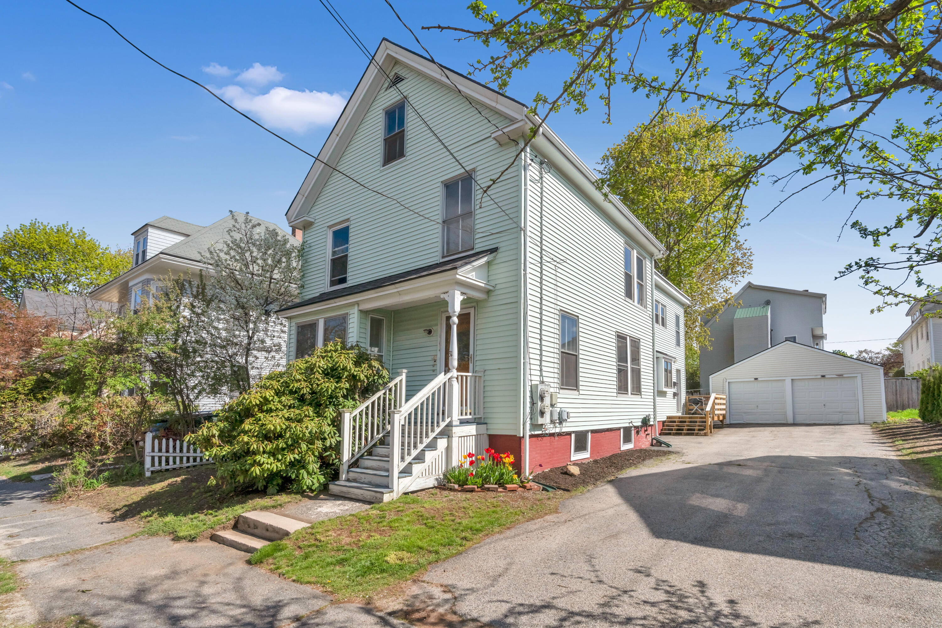 Pale green two-story house with a covered porch, spring flowers, and a detached two-car garage.