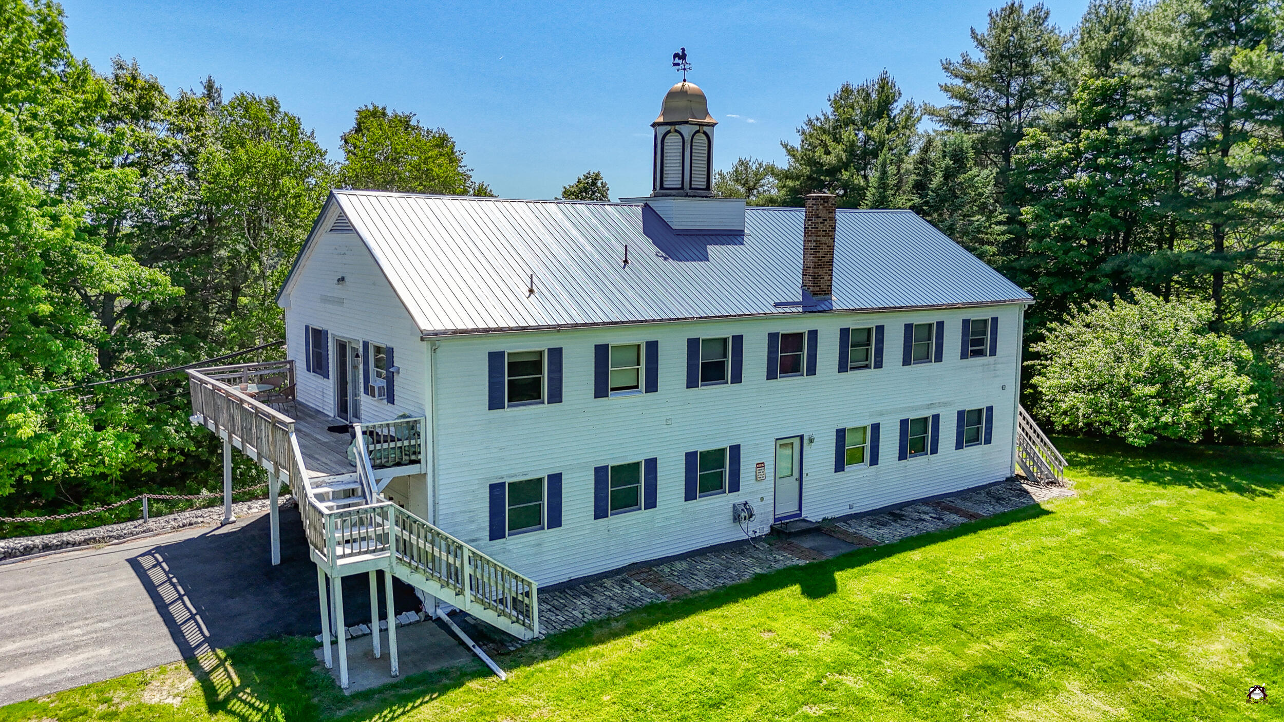 Two-story white building with a metal roof, cupola, and elevated wooden decks amid trees.