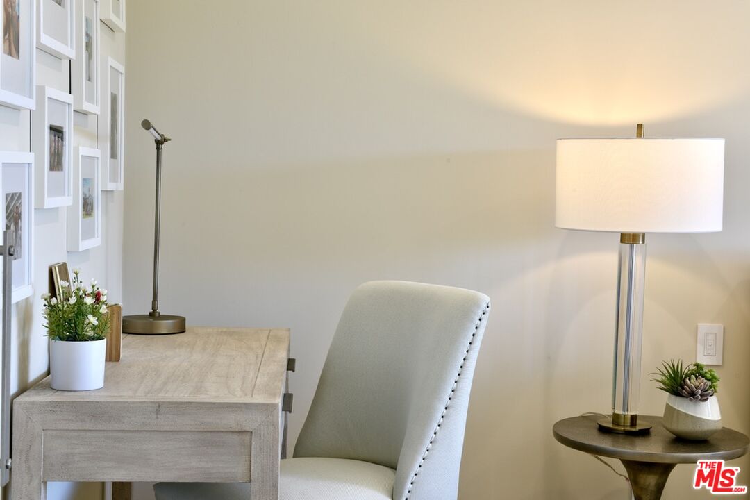 A light-filled desk nook with a white upholstered chair, framed photos, and a tall floor lamp.
