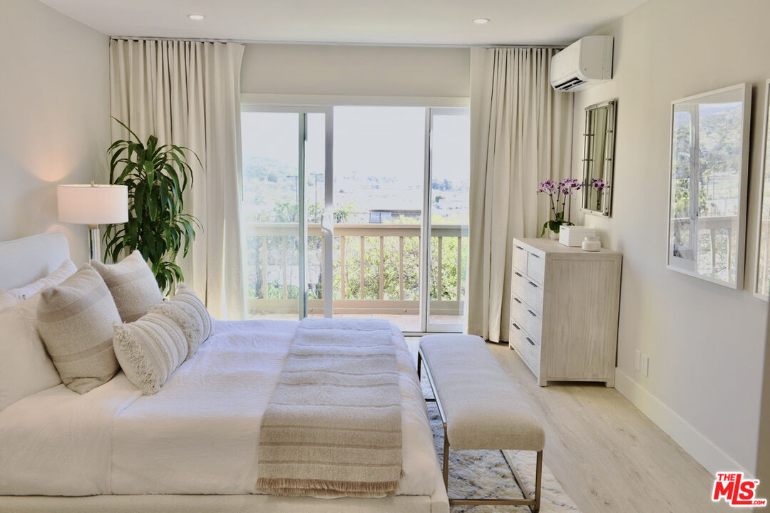 Bright staged bedroom with a white bed, dresser, and sliding glass doors opening to a balcony.