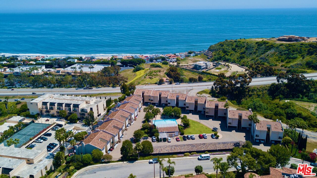 Aerial view of coastal condominium complex with a pool, tennis courts, and oceanfront nearby.