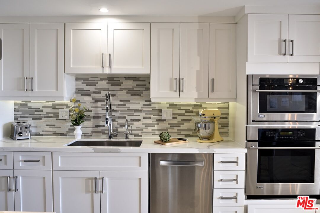 Bright white kitchen with a gray tile backsplash, stainless steel appliances, and a yellow stand mixer.